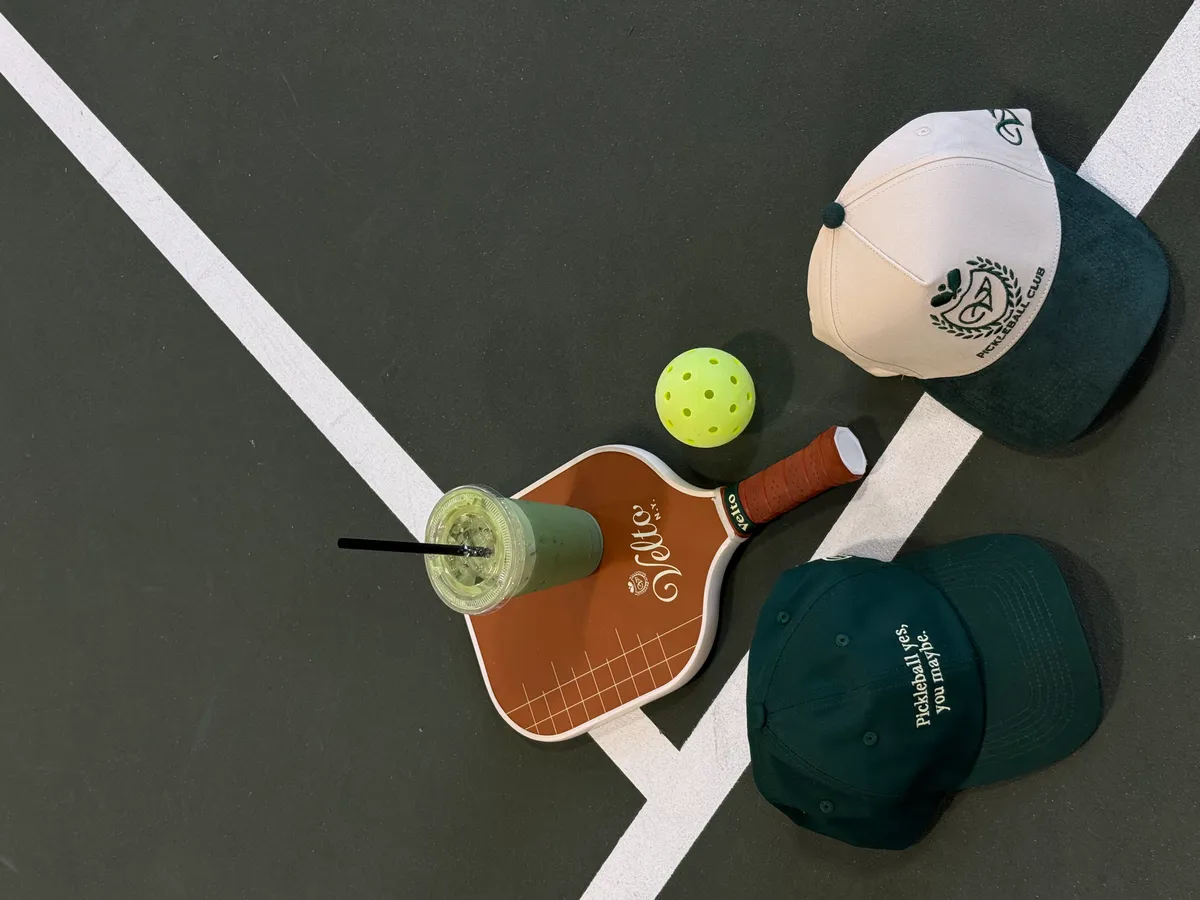 Velto pickleball paddle, ball, iced matcha and two club caps arranged on an indoor court